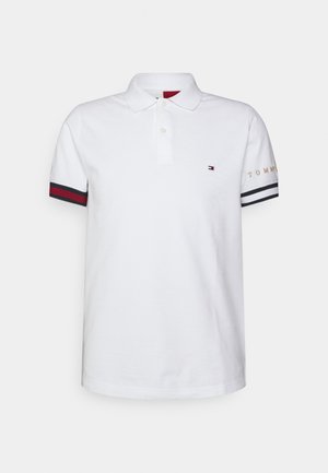 Polo blanc à manches courtes avec patte à deux boutons, logo Tommy Hilfiger sur la poitrine, et poignets rayés avec le texte "TOMMY" sur la manche droite.