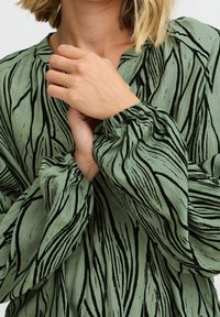 Blouse à motifs verts avec des rayures noires abstraites, dotée d'une coupe décontractée, de manches froncées et d'un col rond. Tissu texturé visible.