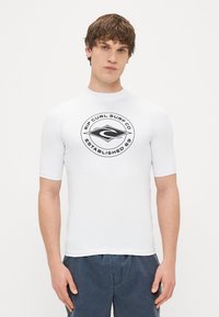 Biała koszulka rash guard z krótkim rękawem, z czarnym okrągłym logo z napisem "Rip Curl Surf Co Established 69". Gładka tekstura, dopasowany krój.