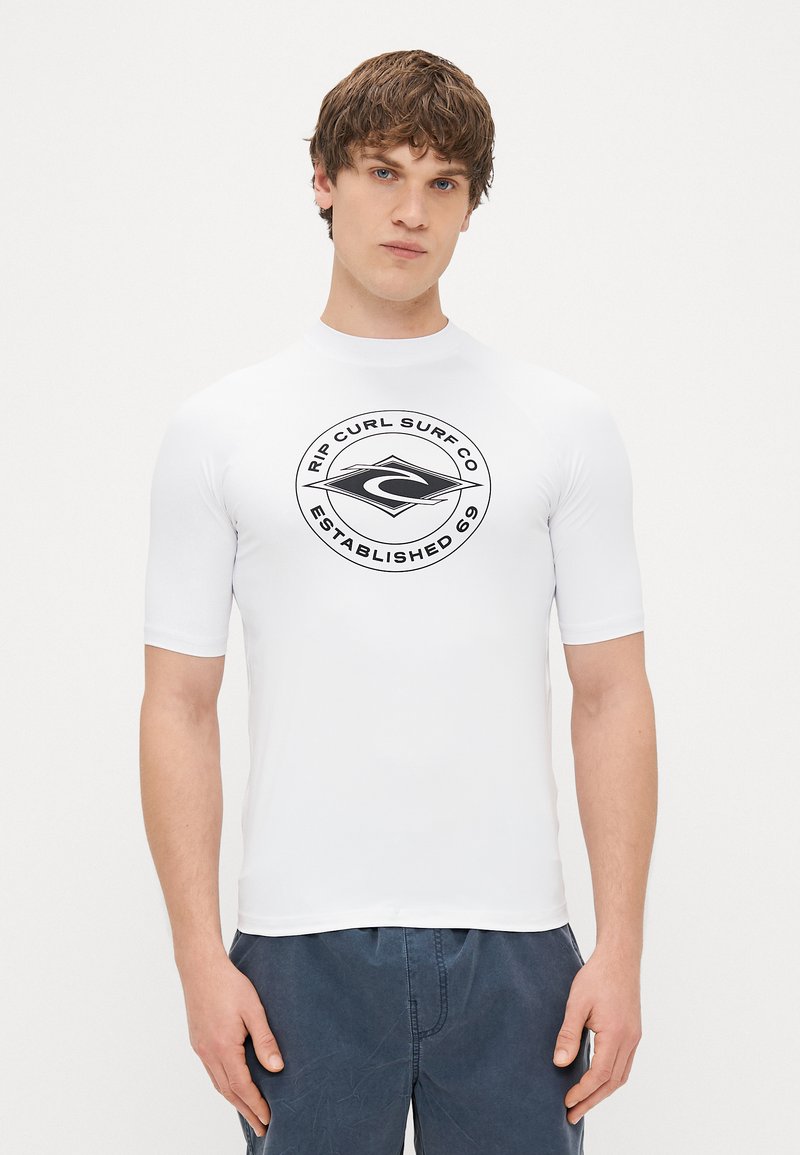 Biała koszulka rash guard z krótkim rękawem, z czarnym okrągłym logo z napisem "Rip Curl Surf Co Established 69". Gładka tekstura, dopasowany krój.