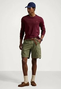 Jovem vestindo uma camisa de manga longa bordeaux, calções cargo verde-oliva com um cinto trançado, meias bege, sapatos castanhos e um boné azul-marinho.