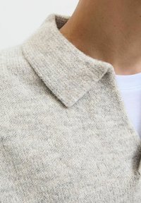 Strikket sweater i lys beige med krave. Blød tekstur med synlig syning. Laget over en hvid skjorte, der fremhæver en afslappet stil.
