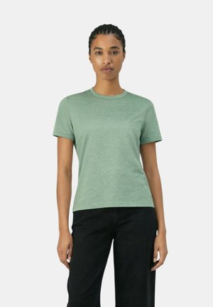 Donna con capelli intrecciati che indossa una t-shirt verde chiaro tinta unita a maniche corte e pantaloni neri, in piedi davanti a uno sfondo bianco.
