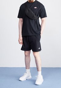 Black T-shirt and shorts, white Nike logo on shorts. Black crossbody bag. White sneakers with visible air sole. Plain background.