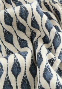 Tessuto a motivi con una base crema e forme di foglie blu scuro disposte in un design ripetuto. Il materiale appare texturizzato e tessuto.