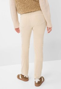 Person, der beiges, gerade geschnittenes Hosen trägt, eine sandfarbene, strukturierte Weste, ein hellbeiges langärmeliges Oberteil und braune Sneakers mit weißen Akzenten, steht mit dem Rücken zur Kamera.