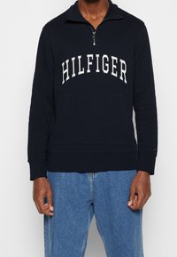 Homme portant un sweat-shirt à fermeture éclair quart de zip bleu marine avec le texte "HILFIGER" sur la poitrine, associé à un jean bleu clair.