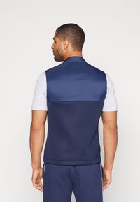 Colete azul-marinho com uma parte superior lisa e brilhante e uma parte inferior texturizada. Apresenta um colarinho levantado e um design ajustado.