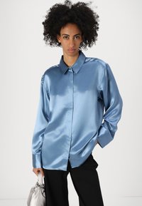 Camicia lunga in satin blu con colletto a punta, chiusura con bottoni e vestibilità leggermente ampia. Il modello tiene una borsa argento.