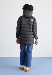 Svart puffy parka jakke med horisontal quiltedesign, hette og logo detalj på baksiden, kombinert med vide jeans og fargerike joggesko.