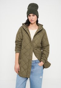 Πράσινο ελιάς μπουφάν με καπιτονέ ύφασμα και κουκούλα, με κουμπιά με τρούκ και ριμπ μανσέτες. Φορεμένο πάνω από ένα ανοιχτό μπεζ τοπ και συνδυασμένο με ανοιχτό μπλε τζιν.