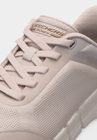 Κοντινό πλάνο μπεζ αθλητικού παπουτσιού Skechers BOBS που δείχνει υφασμάτινη υφή, κορδόνια και ετικέτα με το λογότυπο στη γλώσσα.