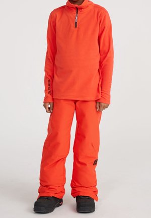 Niño con chaqueta polar naranja brillante con cremallera y pantalones de nieve naranja a juego, con botas de invierno negras, sobre fondo liso.