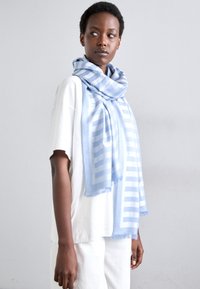 Écharpe bleu clair avec des rayures diagonales, texture douce, bords frangés, enroulée autour du cou, associée à un T-shirt blanc et un pantalon clair.