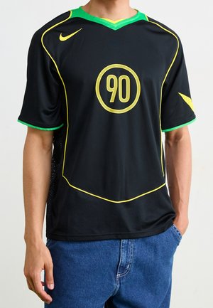 Person wearing a black sports shirt with yellow trim, green collar, yellow number 90 on front, and blue jeans, standing with one hand in pocket.