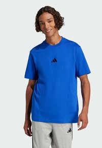 Persona sorridente con capelli ricci che indossa una maglietta blu Adidas e pantaloni corti grigi chiari Adidas su uno sfondo chiaro e uniforme.
