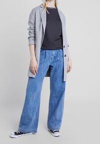 Grå ullblazer över en svart skjorta, med blå vida denimjeans och svart-vita sneakers, med en avslappnad, lös passform.