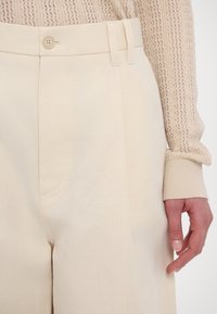 Pantalon beige clair taille haute avec bouton et passants de ceinture, porté avec un pull beige texturé manches longues, montrant une main détendue sur le côté.