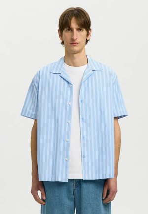 Junger Mann trägt ein hellblau-weiß vertikal gestreiftes kurzärmeliges Hemd über einem weißen T-Shirt und blauen Jeans, steht neutral.