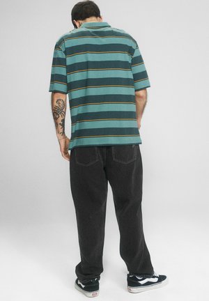 Mężczyzna zwrócony tyłem, ubrany w zielononiebieską, prążkowaną koszulkę z krótkim rękawem, czarne spodnie, czarno-białe trampki, z tatuażami na lewym przedramieniu.