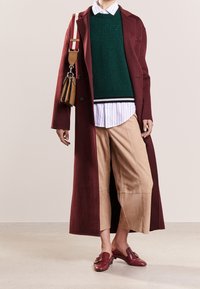Long manteau bordeaux sur un pull en laine vert, chemise crème à rayures, pantalon beige court et chaussures à enfiler bordeaux avec des détails en métal.