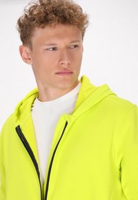 Heldere gele zip-up hoodie met zwarte rits, lichtgewicht stof en een aangehechte capuchon, gedragen over een wit shirt. Gladde textuur.