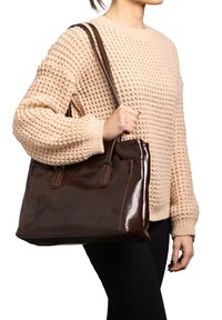 Bruine leren handtas met een gladde afwerking, dubbele handvatten en een gestructureerde vorm. Wordt vastgehouden door een persoon die een textuurcrème trui draagt.