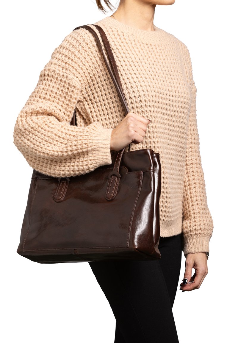 Bruine leren handtas met een gladde afwerking, dubbele handvatten en een gestructureerde vorm. Wordt vastgehouden door een persoon die een textuurcrème trui draagt.