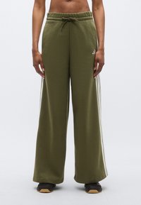 Zielone spodnie dresowe o szerokich nogawkach z regulowanym paskiem i białymi paskami po bokach. Materiał wydaje się miękki i gładki w dotyku.