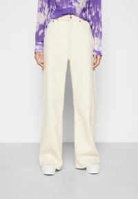 Calças de perna larga em algodão creme com um acabamento texturizado. Apresenta uma cintura alta, design de cinco bolsos e fecho frontal com botão.