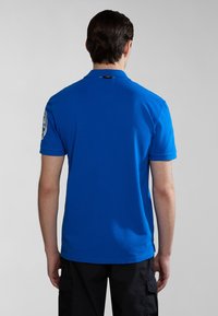 Modrá polo tričko s krátk�ým rukávem, límcem a logem na levém rukávu. Hladká textura, přizpůsobený střih a minimalistický design.
