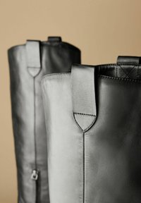 Las botas de cuero negras tienen la parte superior redondeada, costuras fuertes y tiradores. La superficie es suave, con detalles de costura y una cremallera lateral.
