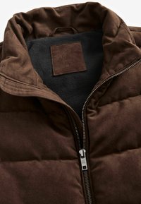 Brun puffer vest med lynlås foran, blødt indre for og et rektangulært mærke indeni. Tekstureret ydre med kviltet design.