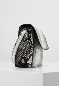 Černobílá kabelka ve stylu hadí kůže se strukturovaným tvarem, hladkým povrchem a klopovým uzávěrem. Doplněna tenkým popruhem a lesklým povrchem.