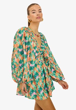 Femme portant une robe courte à fleurs colorées avec des manches longues bouffantes, un décolleté en V profond et une taille cintrée, accessoirisée de boucles d'oreilles créoles dorées et d'un collier.