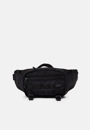 Marsupio in tessuto nero con tasca frontale con zip, spallacci regolabili e piccolo logo Nike davanti su sfondo bianco.