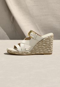 Sandale compensée blanche avec talon en corde tressée, boucle dorée et brides croisées sur une surface beige avec un fond en tissu doux.