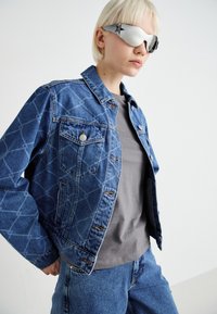 Denim jakna z geometrijskim vzorcem, modre barve, z gumbastim zapiranjem, dvema sprednjima žepoma, v kombinaciji s sivo majico in usklajenimi modrimi kavbojkami.