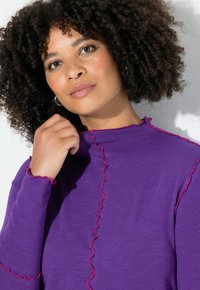 Lila långärmad topp med hög halsringning, med kontrasterande röd rynkad söm längs sömmarna och ärmslut, tillverkad av mjukt tyg.