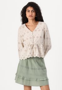 Beige eyelet blouse with a V-neck, long sleeves, and drawstring waist. Paired with a layered green skirt featuring ruffles and lace accents.