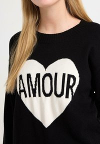 Czarny sweter z dużym białym graficznym sercem oraz słowem "AMOUR" napisanym wyraźnymi czarnymi literami. Żebrowany dekolt i miękka tekstura.