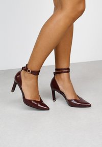 Zapatos de tacón alto burdeos con un diseño de punta afilada. Cuentan con una correa en el tobillo con hebilla y una textura de cuero brillante.