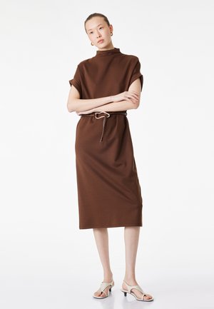 Donna in piedi con le braccia incrociate, indossa un vestito midi marrone con maniche risvoltate, una sottile cintura, orecchini a cerchio dorati e tacchi beige aperti in punta.