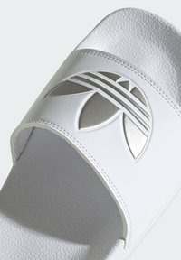 White slide sandal with textured synthetic upper, featuring a silver embossed logo and stitching details. Open-toe design with smooth finish.