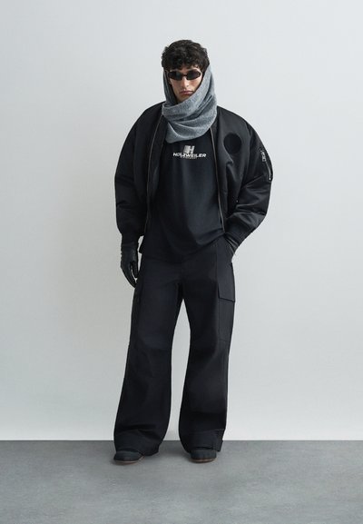 Μαύρο bomber μπουφάν με υφασμάτινο γκρι κασκόλ, μαύρο T-shirt με λογότυπο, φαρδιά μαύρα cargo παντελόνια, μαύρα γάντια και γυαλιά ηλίου.