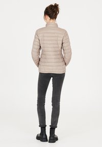 Beige puffer jacket met verticale gestikte patroon, hoge kraag en lichtgewicht materiaal. Gecombineerd met donkergrijze skinny jeans en zwarte enkellaarsjes.