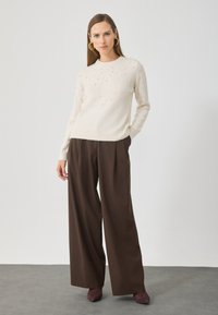 Světle krémový pletený svetr s perlovými ozdobami, v kombinaci s tmavě hnědými kalhotami s širokými nohavicemi a burgundskými podpatky na šedé podlaze.