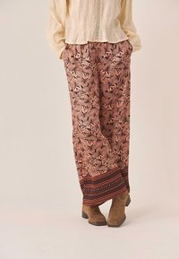 Pantalones fluidos y estampados en tonos de rosa y marrón con diseños florales y una banda decorativa trenzada en el dobladillo, combinados con botas de tobillo marrones.