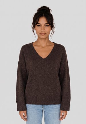 Donna con capelli scuri raccolti in uno chignon, indossa un maglione marrone scuro a scollo a V e jeans azzurri chiari, in piedi davanti a uno sfondo grigio chiaro uniforme.