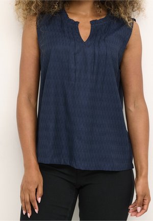 Kobieta w granatowej bluzce bez rękawów z małym wycięciem w kształcie litery V i czarnych spodniach, stojąca z ramionami swobodnie opuszczonymi wzdłuż ciała.